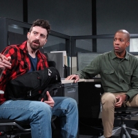 Two men sitting at a desk, one pointing away and the other looking at him.