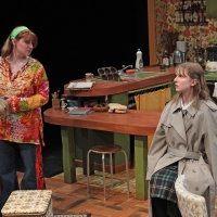 Young woman in an orange shirt looking askance at a young woman sitting at a table.