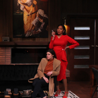 A woman sitting on a couch, another woman standing behind her, both in front of a painting of two women