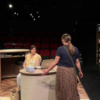 A woman in a yellow top sitting at a desk facing off against a woman standing on the other side of her