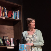 A woman standing in front of a book case looking away, holding a book.
