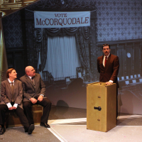 Two men sitting watching another man speaking at a podium