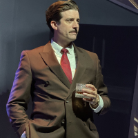 A man in a 1930s suit and pencil mustache standing and looking off