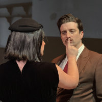 A woman dressed in black putting her finger on a man's mouth to stop him from speaking