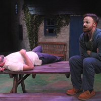 One man lying on a bench, another man sitting on a bench next to him