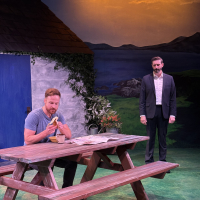 One man sitting at a picnic table and another standing behind him looking at him