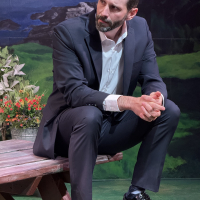 A bearded man in a suit sitting on a picnic table holding his hands together and looking off to the left