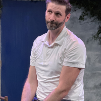 A bearded man in a white polo shirt sitting on a table and looking ahead